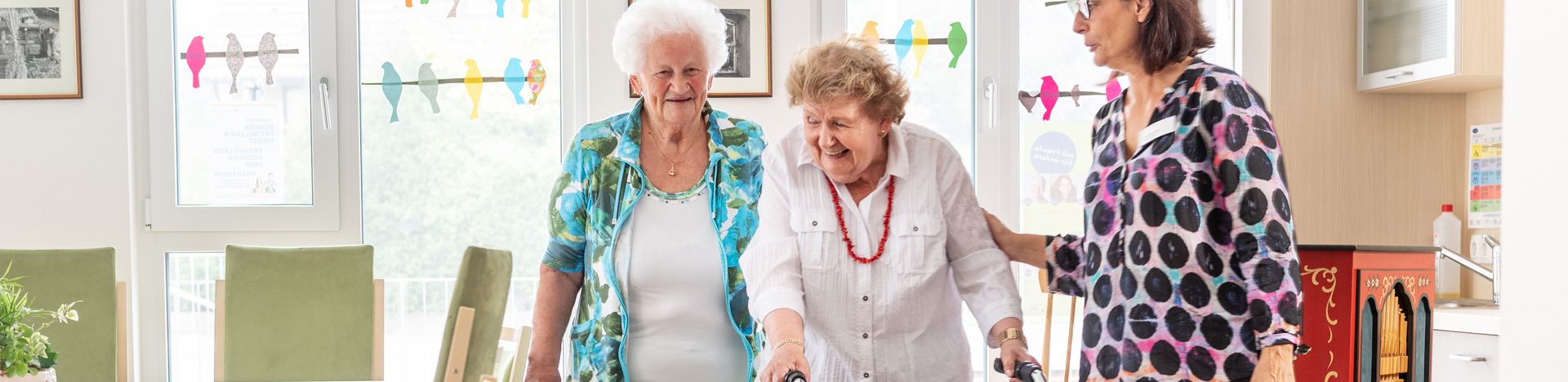 Zwei Frauen unterstützen ältere Frau beim Gehen mit Rollator